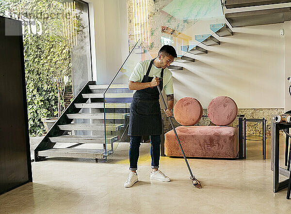 Young man wearing apron cleaning floor with broom in cafe