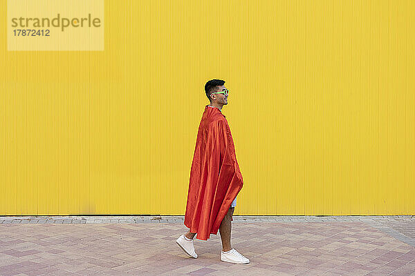Young man wearing red superhero cape walking in front of yellow wall