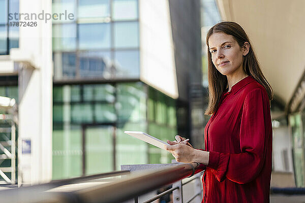 Confident businesswoman with tablet PC by railing at workplace