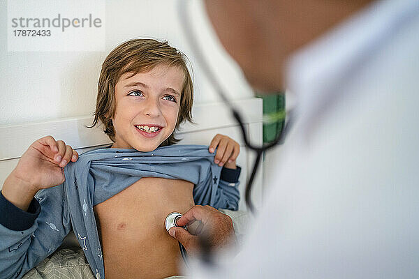 Doctor with stethoscope examining smiling boy in bed at home