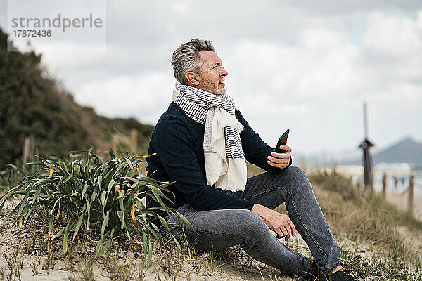 Reifer Mann mit Smartphone sitzt neben einer Pflanze am Strand