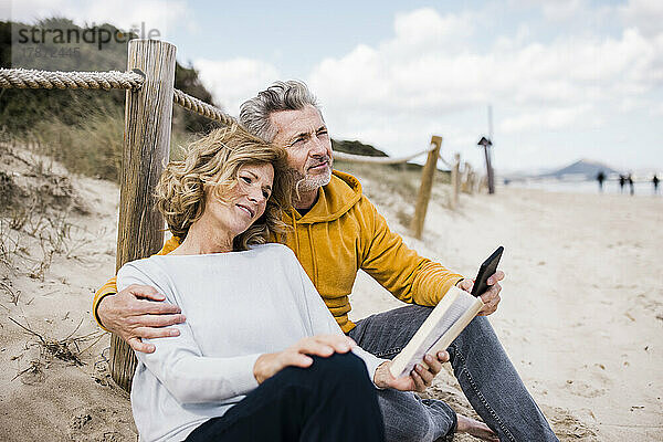 Lächelnder reifer Mann mit Frau  die am Strand ein Buch liest