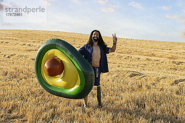 Lächelnder Mann mit Beinprothese  der einen avocadoförmigen aufblasbaren Ring hält und auf dem Feld ein Friedenszeichen zeigt