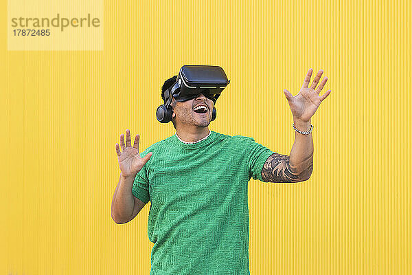 Glücklicher junger Mann mit Virtual-Reality-Simulator gestikuliert vor gelbem Hintergrund