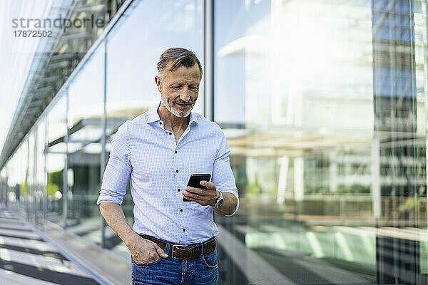 Reifer Geschäftsmann mit der Hand in der Tasche und Smartphone an der Glaswand