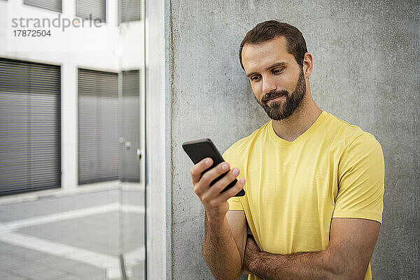 Junger junger Mann mit Bart benutzt Mobiltelefon und steht vor der Wand