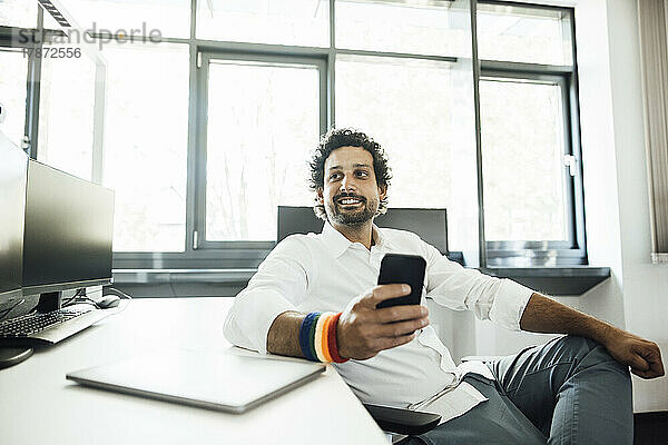 Lächelnder Geschäftsmann mit Smartphone am Schreibtisch im Büro