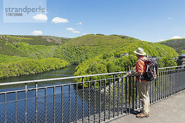 Älterer Mann mit Hut und Rucksack steht am Geländer und blickt auf die Aussicht  Nationalpark Eifel  Deutschland