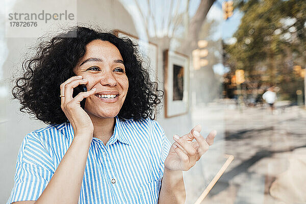 Glückliche Frau gestikuliert und spricht mit dem Mobiltelefon  gesehen durch Glas im Café