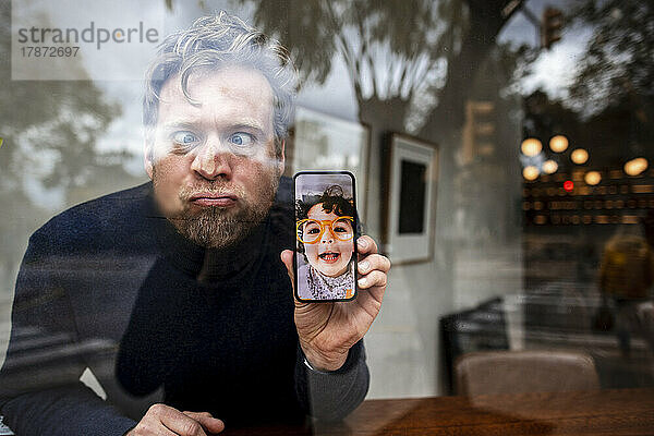 Verspielter Geschäftsmann mit gekreuzten Augen zeigt das Gesicht seiner Tochter bei einem Videoanruf im Café