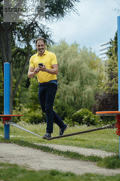 Glücklicher Mann balanciert mit Smartphone auf Seil im Park