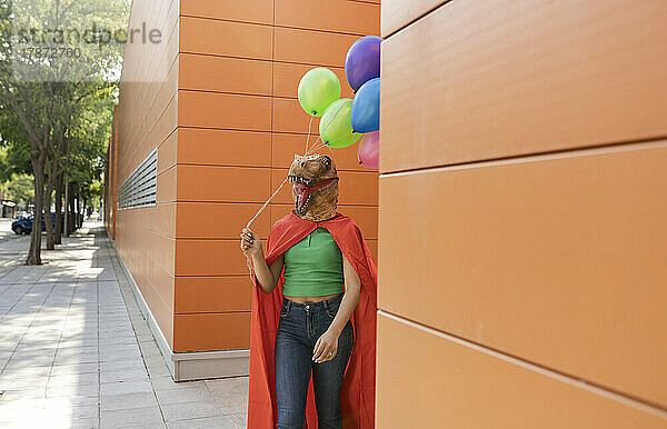 Frau mit Krokodilmaske läuft mit bunten Luftballons vor der Wand