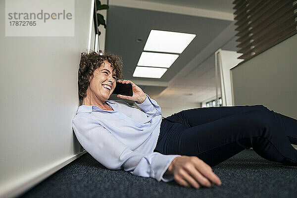 Cheerful businesswoman talking on smart phone reclining in office