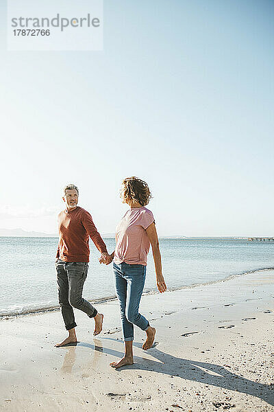 Reifer Mann und Frau halten sich an den Händen und gehen am Strand spazieren