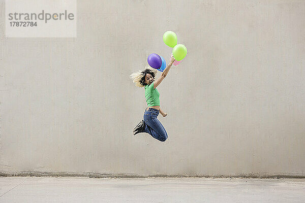 Glückliche junge Frau mit bunten Luftballons  die vor der Wand springt