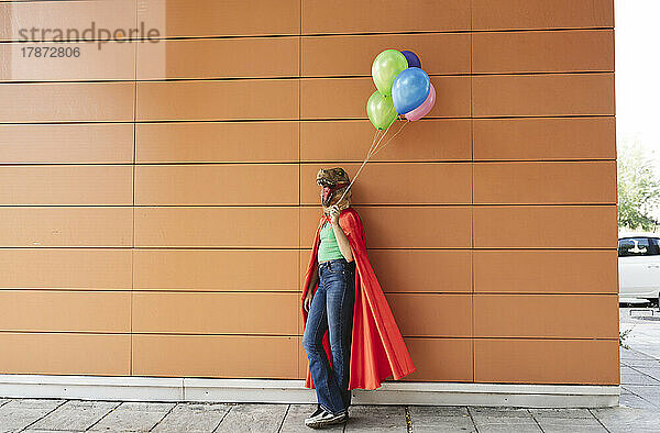 Frau mit Krokodilmaske lehnt mit bunten Luftballons an der Wand