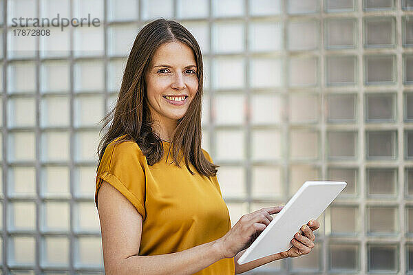 Smiling businesswoman with tablet PC by wall in office