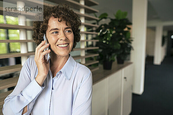 Glückliche Geschäftsfrau mit lockigem Haar  die im Büro mit dem Handy spricht