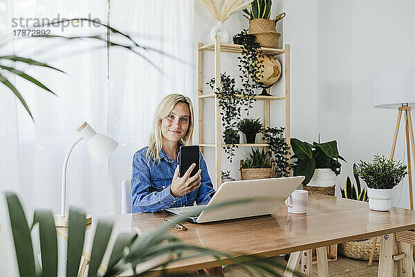 Lächelnder Freiberufler mit Smartphone und Laptop im Heimbüro