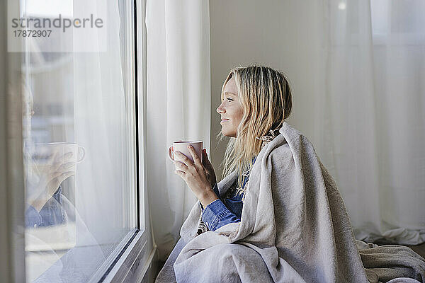 Lächelnde Frau  in eine Decke gehüllt  hält eine Tasse in der Hand und schaut zu Hause aus dem Fenster