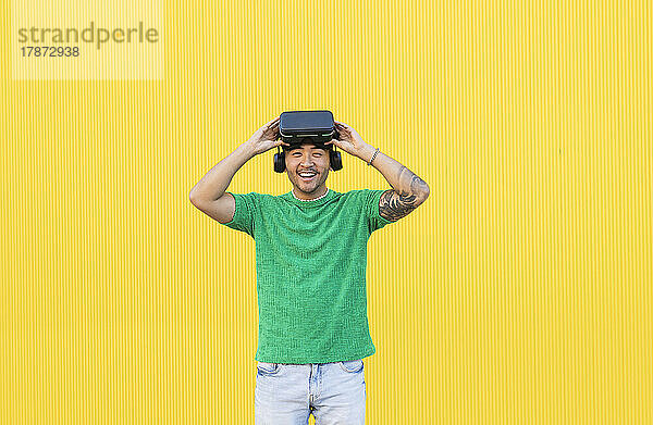 Glücklicher junger Mann steht mit Virtual-Reality-Simulator vor gelbem Hintergrund