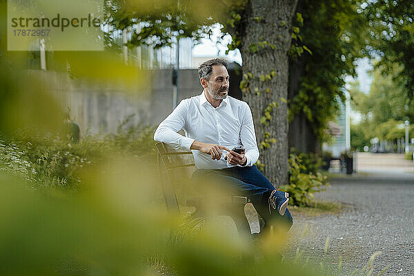 Reifer Geschäftsmann mit Smartphone sitzt auf Bank im Park