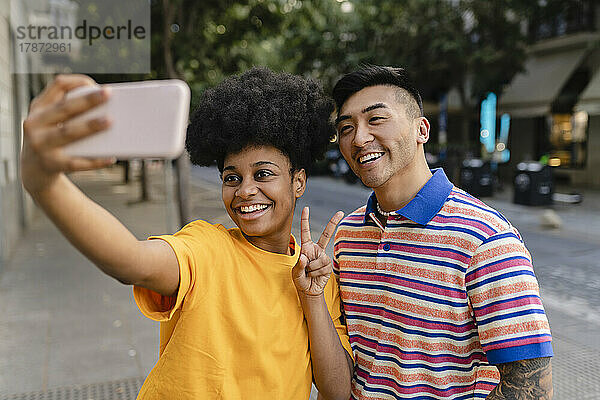 Glückliche junge Frau gestikuliert ein Friedenszeichen und macht ein Selfie mit dem Handy mit einem Mann