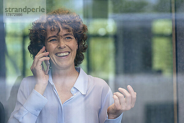 Glückliche Geschäftsfrau  die durch Glas gesehen am Mobiltelefon spricht