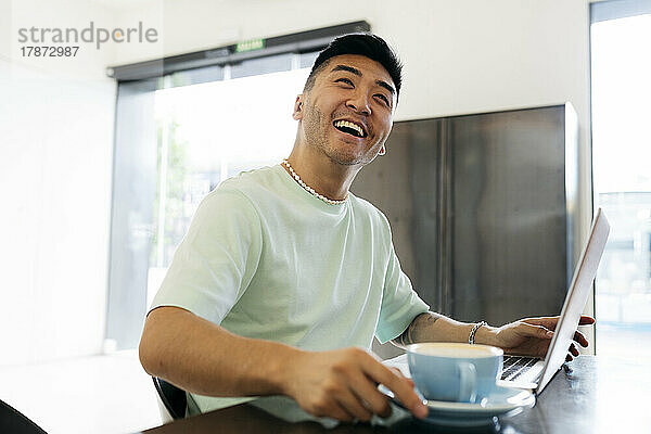 Glücklicher Mann mit Laptop und Kaffeetasse  der am Tisch im Café sitzt
