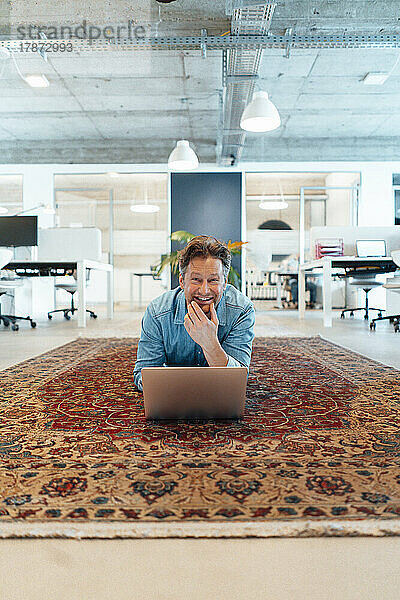 Happy mature businessman lying down with laptop on carpet in office