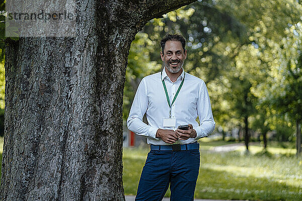 Glücklicher Geschäftsmann hält Smartphone am Baum