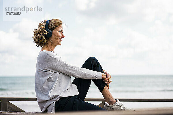 Lächelnde reife Frau mit kabellosen Kopfhörern sitzt am Strand