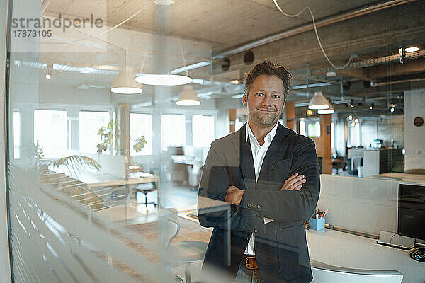 Happy mature businessman standing with arms crossed seen through glass in office