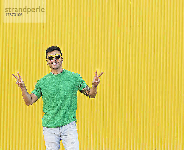 Glücklicher junger Mann gestikuliert Friedenszeichen vor gelber Wand