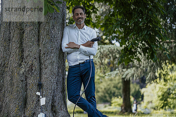 Lächelnder reifer Geschäftsmann mit verschränkten Armen  der das Ladekabel an einen Baumstamm lehnt