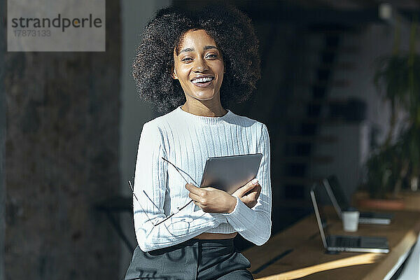Lächelnde Geschäftsfrau mit Brille und Tablet-PC sitzt am Schreibtisch im Büro