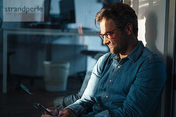 Lächelnder Geschäftsmann mit Brille und Smartphone im Büro sitzend