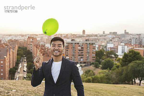 Glücklicher junger Geschäftsmann mit grünem Ballon  der auf einem Hügel steht