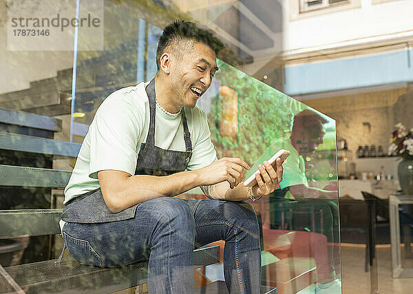 Fröhlicher Mann mit Smartphone sitzt auf Stufen und sieht durch Glas