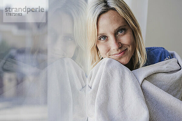 Lächelnde Frau  in eine Decke gehüllt  lehnt an Fensterglas
