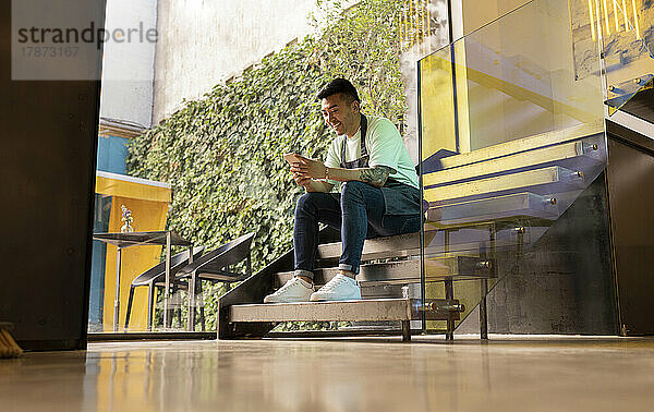 Lächelnder junger Mann  der Textnachrichten über sein Smartphone sendet und auf einer Treppe sitzt