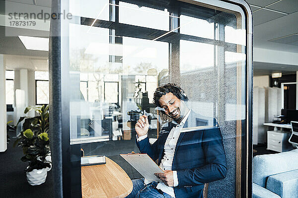 Glücklicher Geschäftsmann mit Klemmbrett  der in einer schallisolierten Kabine im Büro sitzt