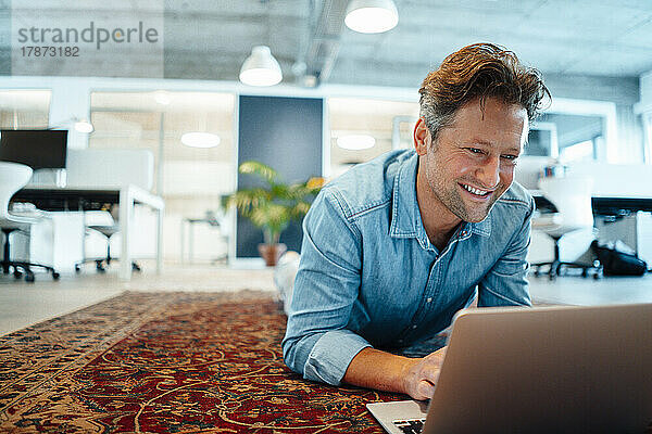 Happy businessman using laptop lying on carpet in office