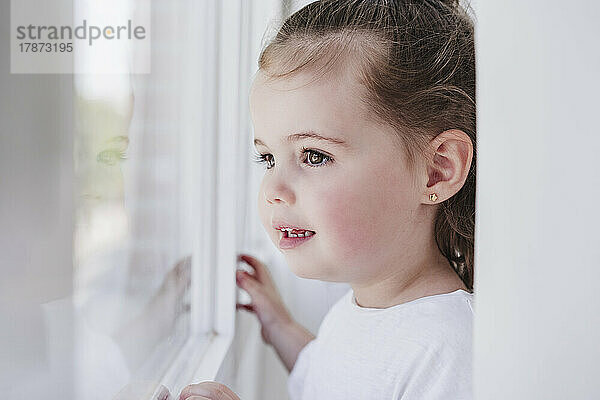 Nachdenkliches süßes Mädchen  das zu Hause aus dem Fenster schaut