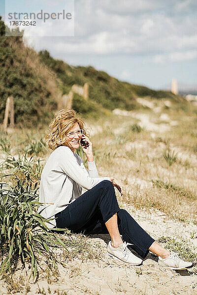 Glückliche reife Frau  die am Strand neben einer Pflanze sitzt und mit ihrem Smartphone spricht