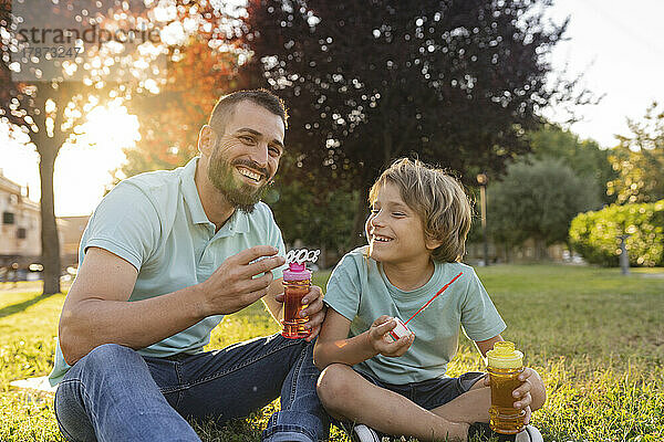Lächelnder Vater und Sohn mit Seifenblasenstäben sitzen im Park