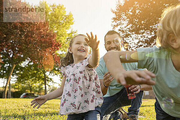 Mann schaut Sohn und Tochter zu  die im Park mit Seifenblasen spielen