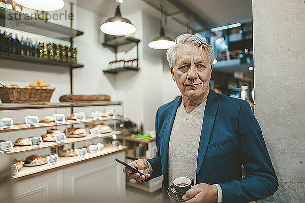 Leitender Geschäftsmann mit Kaffeetasse und Mobiltelefon im Café