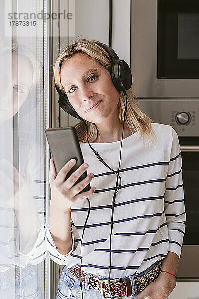 Lächelnde Frau mit Mobiltelefon  die in der heimischen Küche Musik hört