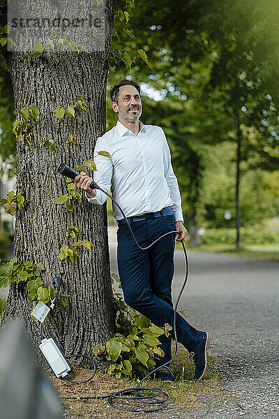 Lächelnder reifer Geschäftsmann hält Ladekabel in der Hand und lehnt an Baumstamm